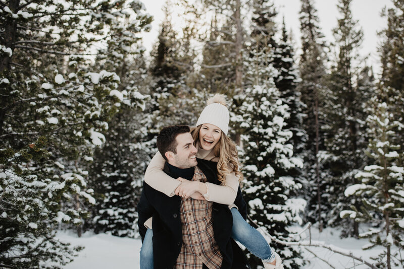 photographers jackson hole photographs an adventurous engagement session in the grand tetons with woman piggy backing an laughing with man in the woods covered in snow