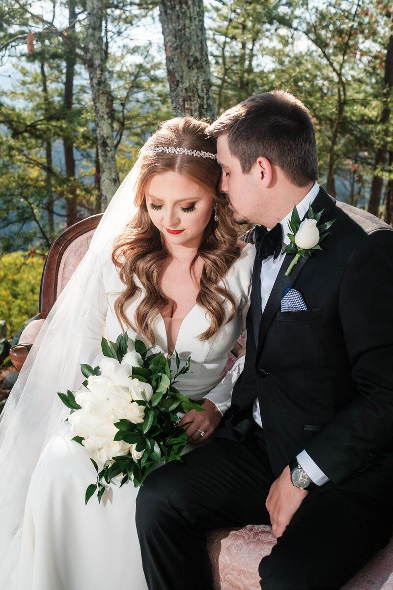 A bride and groom share an intimate moment in a stunning mountain setting, surrounded by lush trees and golden sunlight. The groom leans in to whisper to his bride as she holds a bouquet of elegant white roses. This breathtaking outdoor portrait captures the romance and elegance of Alabama wedding photography.