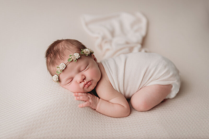 Newborn girl laying on tummy with legs and arms tucked under her and cheek resting on hand wearing a white swaddle and white flower headband