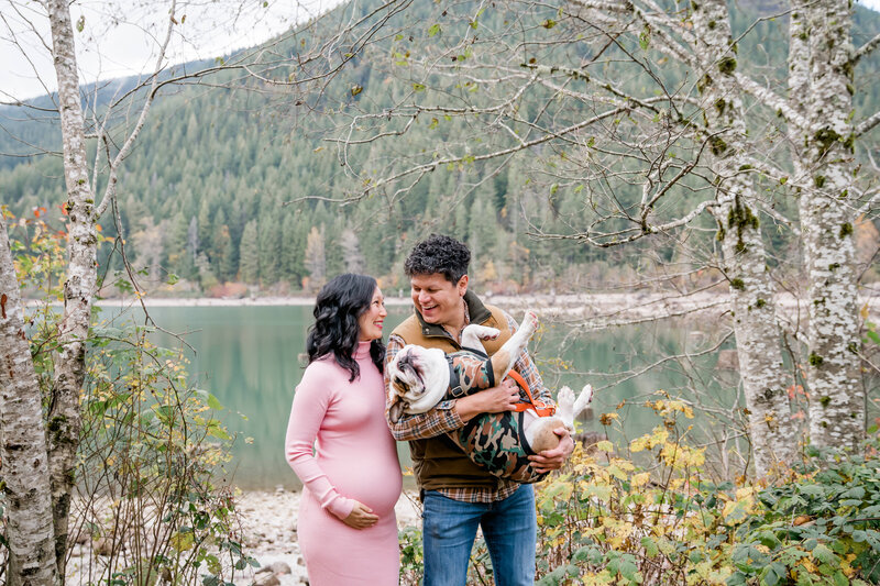 Man and pregnant woman holding a dog standing in front of a lake