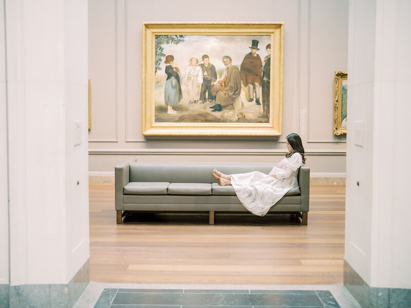 Mom sits on couch in National Gallery of Art with hands on her pregnant belly with her legs up on the couch.