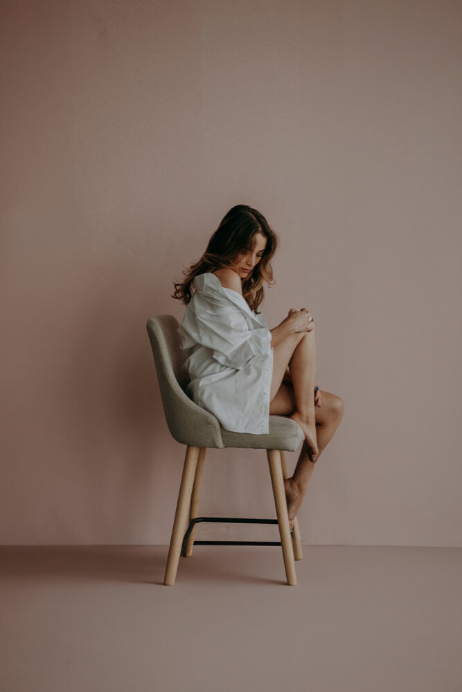Woman sitting on chair in loose button-up shirt, soft pink backdrop, refined and personal boudoir portrait.
