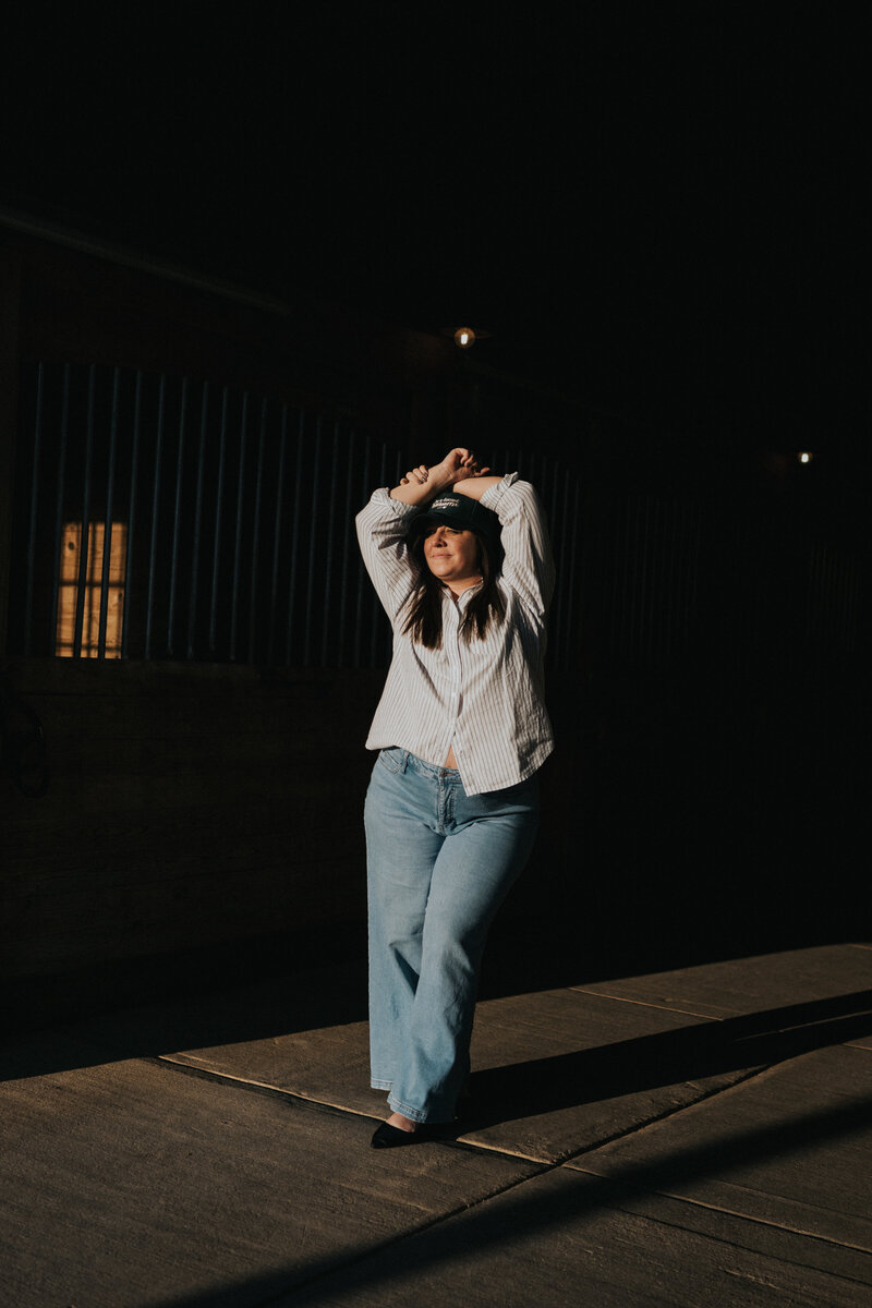 Jo Rickert picture of a midwest photographer and coordinator standing in the light beaming through a barn door on a Nashville horse barn with a "be a decent human" baseball hat  with flared jeans and heels.  entrepreneur headshot boss lady