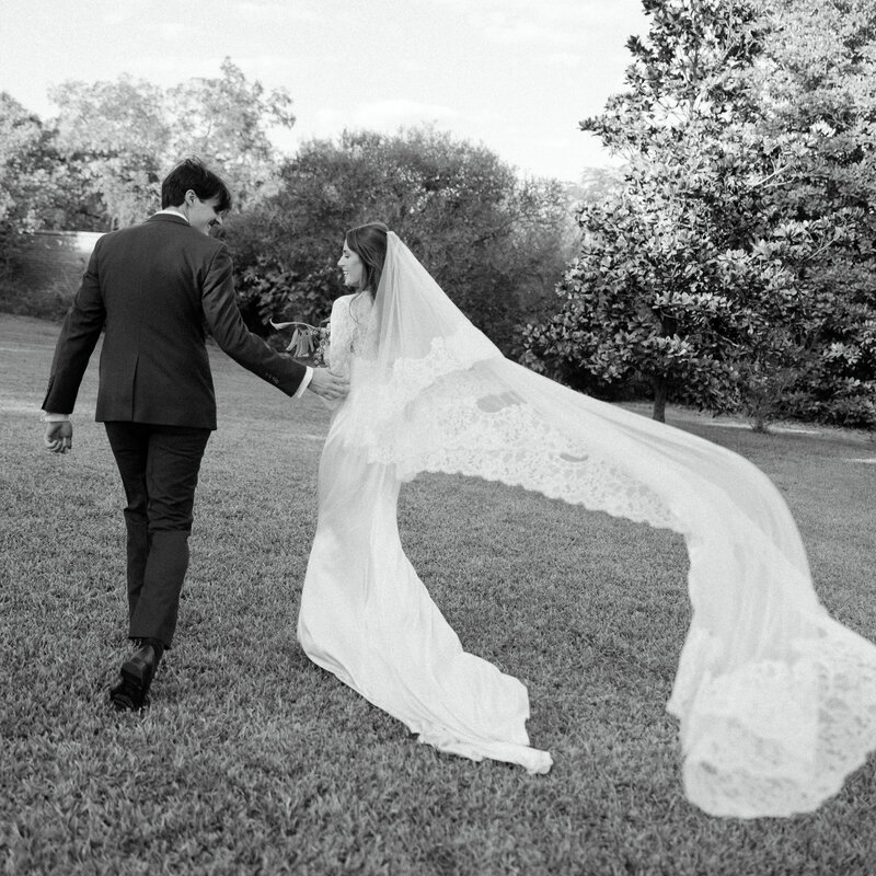 A romantic black and white wedding portrait at Barr Mansion in Austin, Texas, featuring a bride and groom walking hand in hand, with the bride’s lace veil flowing behind her