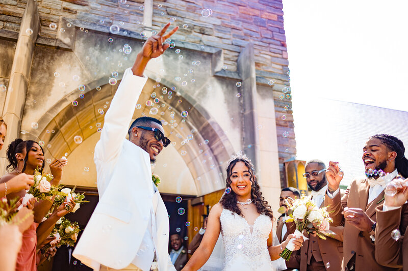 A wedding couple holding hands and laughing as they exit the church after the ceremony in Toledo Ohio. The groom throwing his hand in the air giving the peace sign