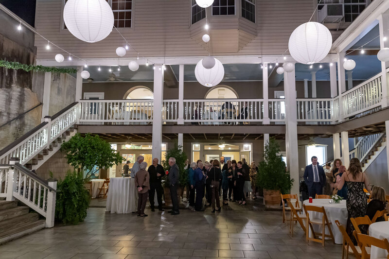 A group of people gathers in a spacious, well-lit courtyard decorated with white paper lanterns and string lights. There are two levels with white railings, scattered tables, and trees adding greenery to the festive scene.