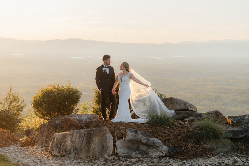 A bridge and groom standing side by side, holding hands and smiling at each other. Behind them are the Blue Ridge Mountains in Morganton NC.