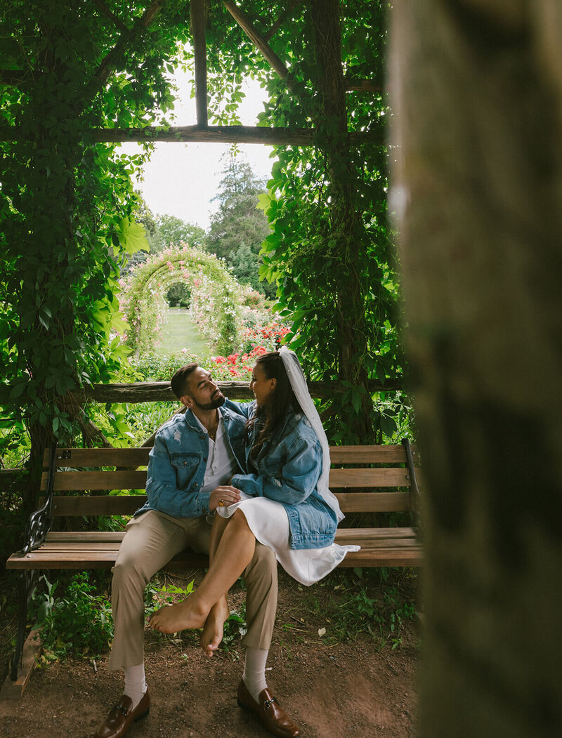 The groom and bride are sitting on a bench in the garden, looking at each other.