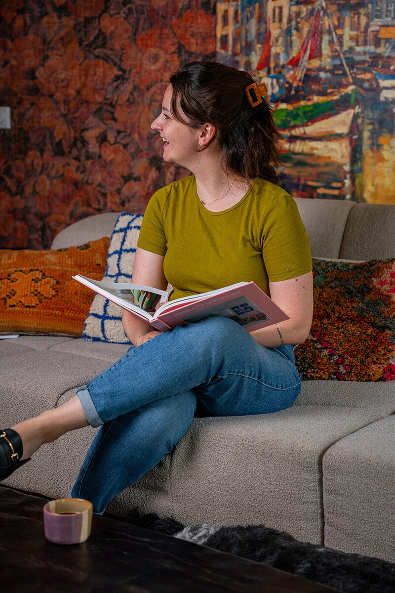Vrouw leest interieurboek op bank met kleurrijk wandkleed en kussens op de achtergrond