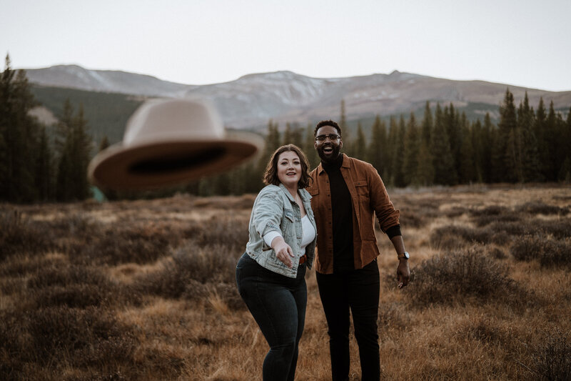 Couple shares playful engagement photo in Breckenridge field, throwing a hat with mountain views in the background.