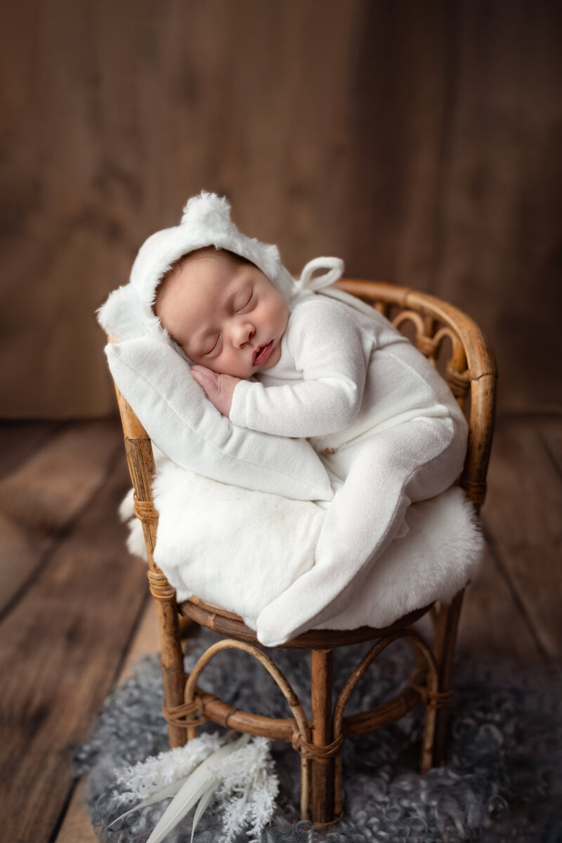 A soft white scene featuring a sleepy newborn boy in a bear bonnet, curled up on a vintage rattan chair.