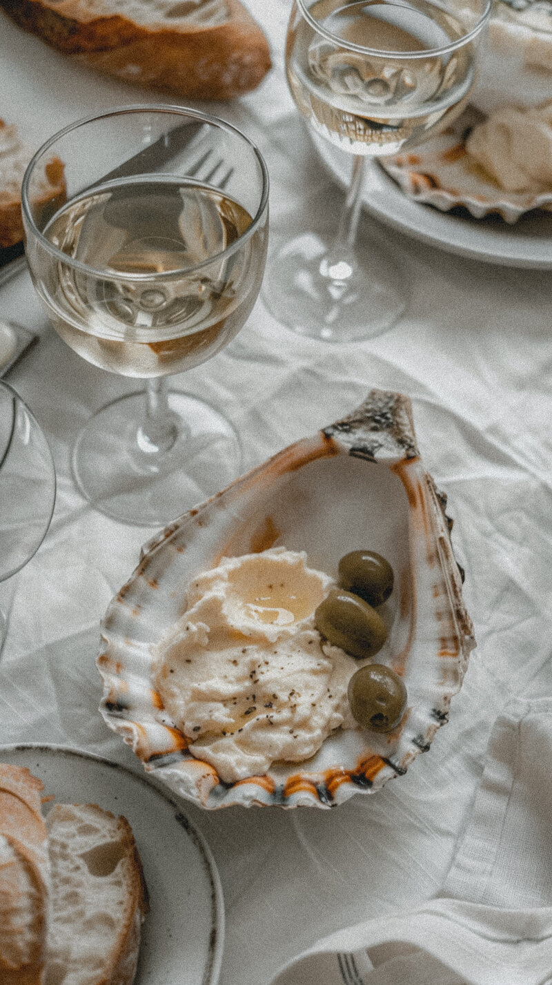 White linen tablecloth large oyster shell with hummus and green olives, a plate of homemade bread slices and wine glasses