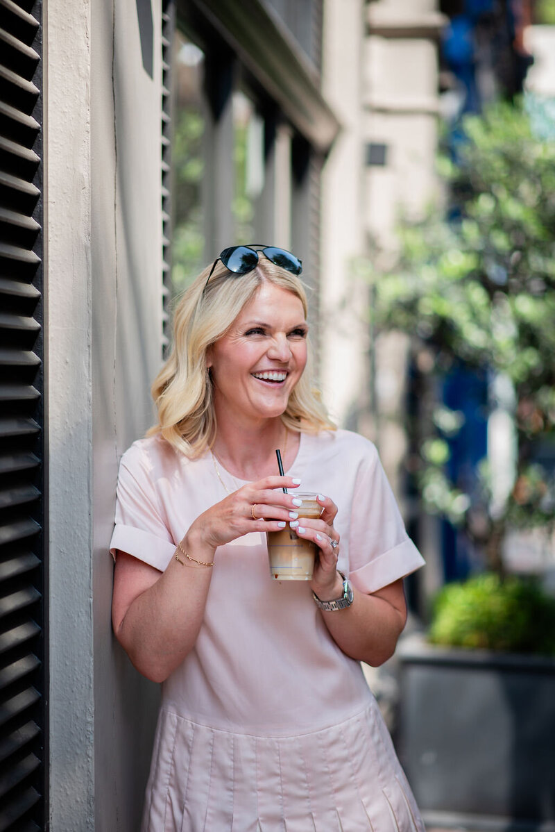 Emily Collins, business coach, smiling and holding a coffee cup while leaning against a brick wall in a black and white photo