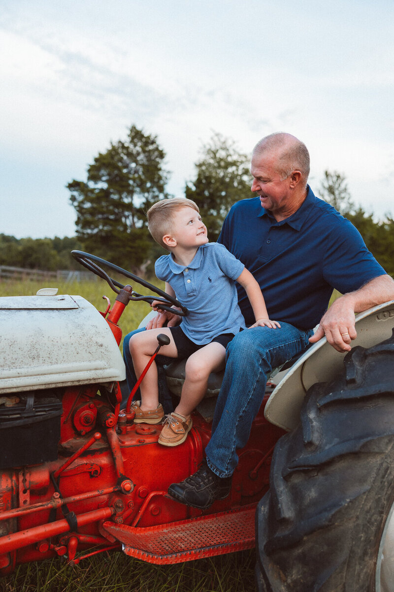 Portrait taken during an extended family session on the family farm of grandpa and his grandson sitting on a red tractor looking at each other. 