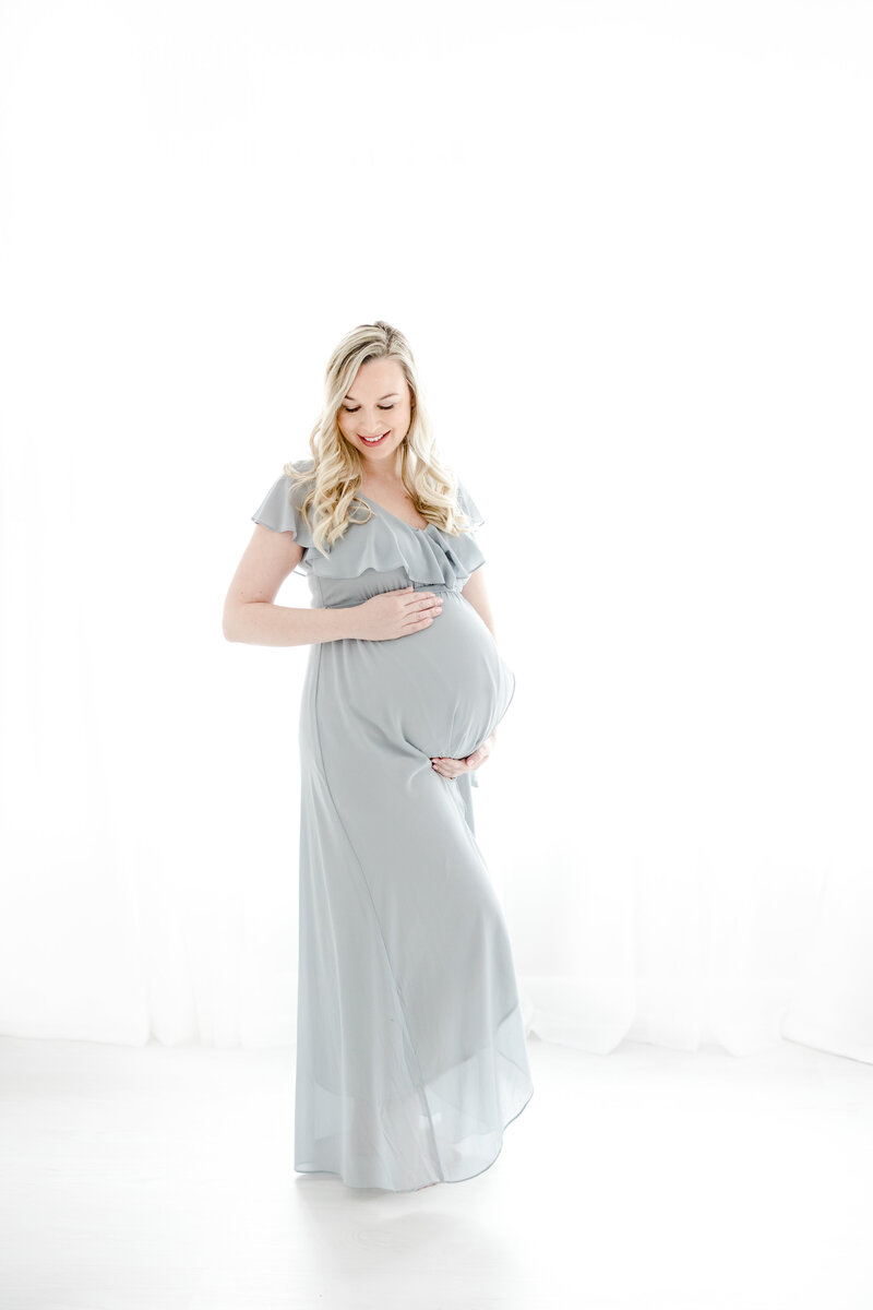 Pregnant woman in light blue flowing gown smiles while holding her pregnant belly during maternity portrait session
