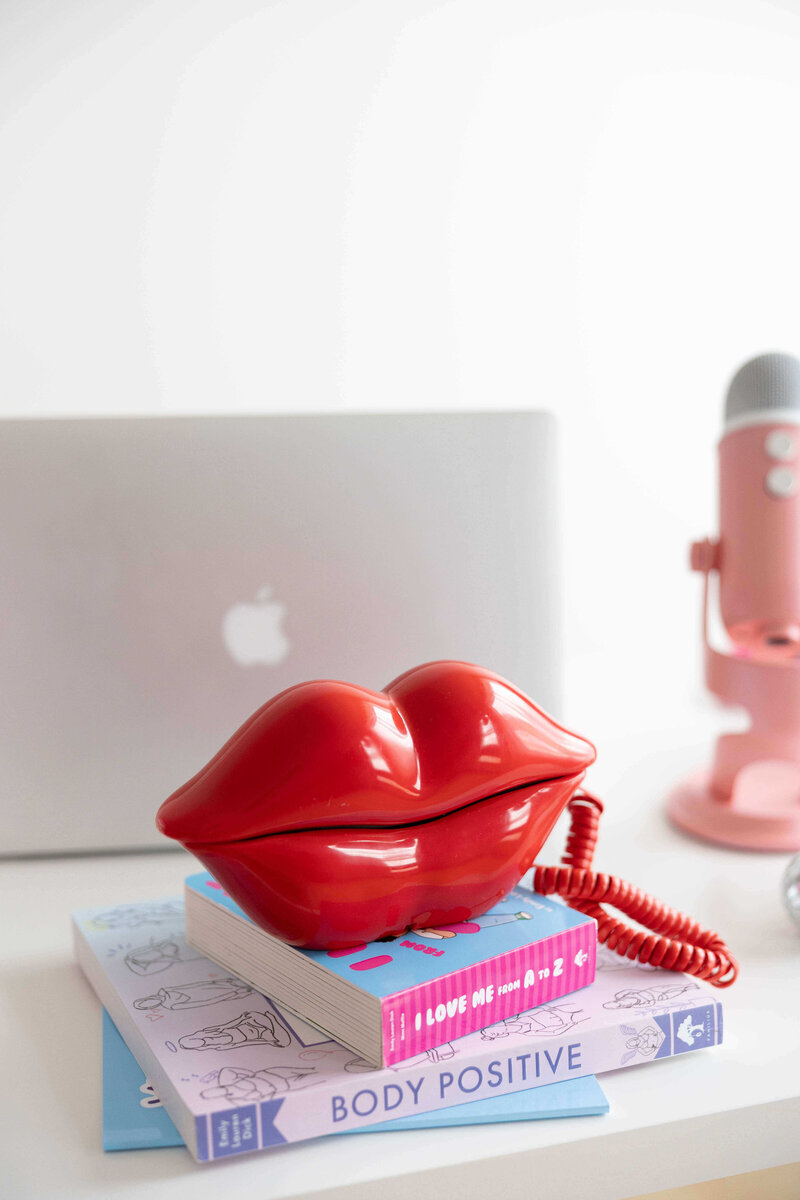 Emily Lauren Dick's books (Body Positive and I Love Me from A to Z) stacked in a pile with a lip shaped phone sitting on top.
