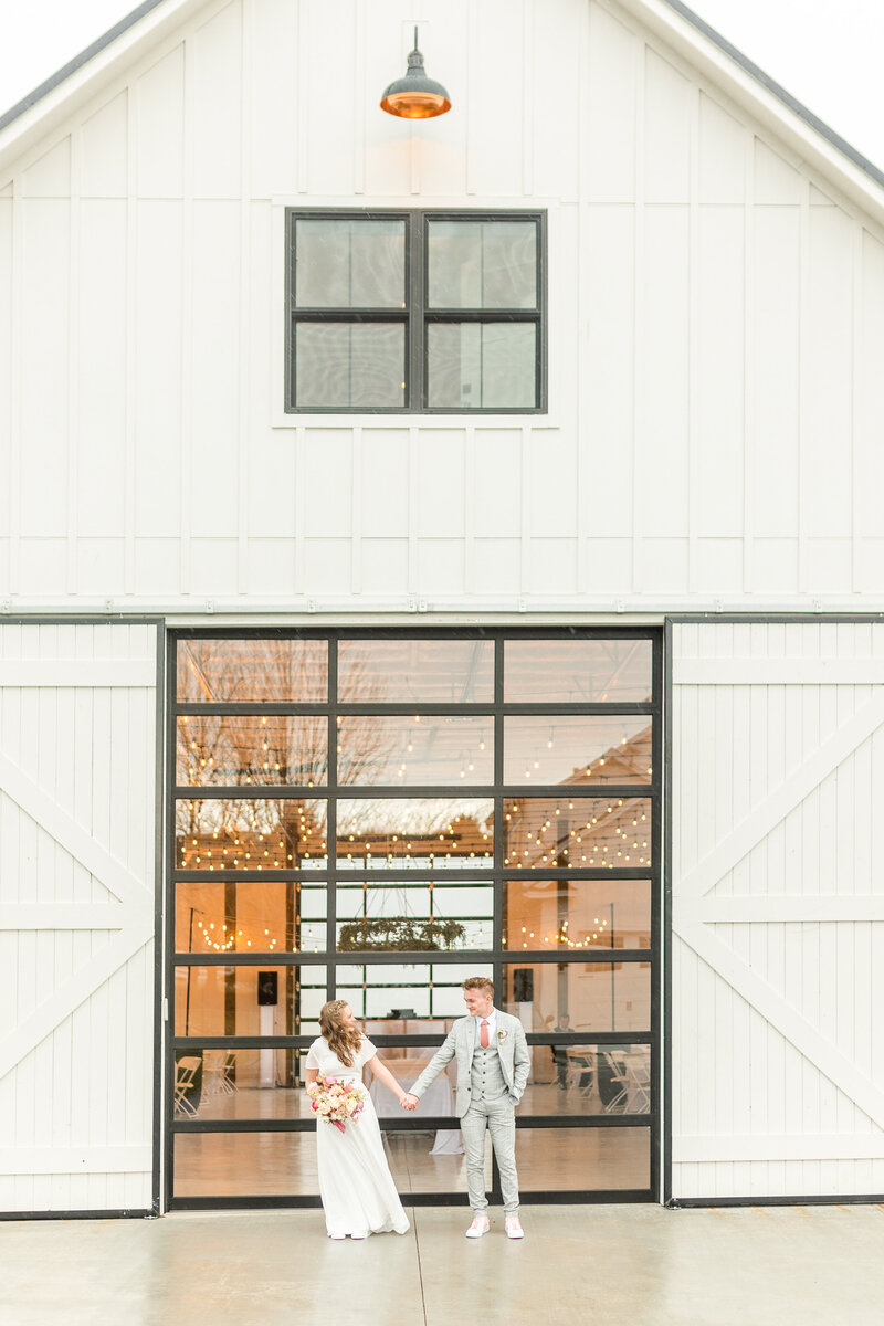 Young couple newlywed holding hands in front of white barn with the inside being totally styled and ready to go