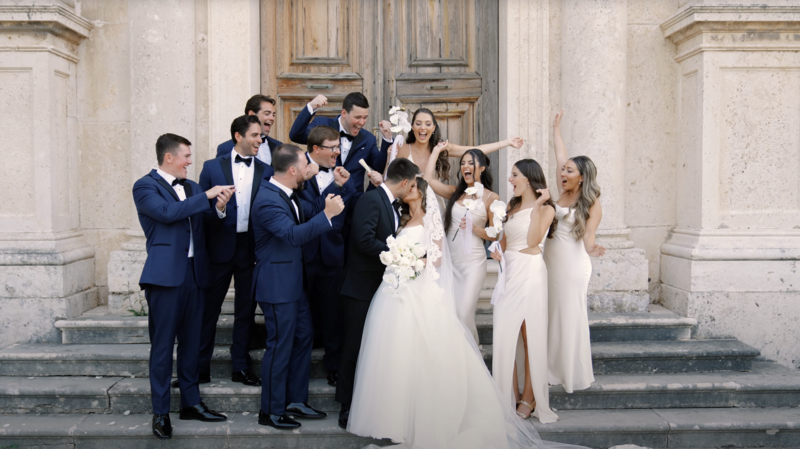 Bride and groom surrounded by joyful bridal party in front of historic stone church in Montenegro, celebrating with laughter and applause — moment from cinematic wedding video abroad