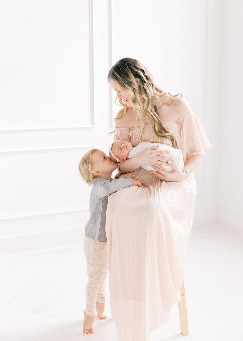 Charlotte family photographer captures a mother holding her newborn baby girl while son kisses little sisters head in a white studio