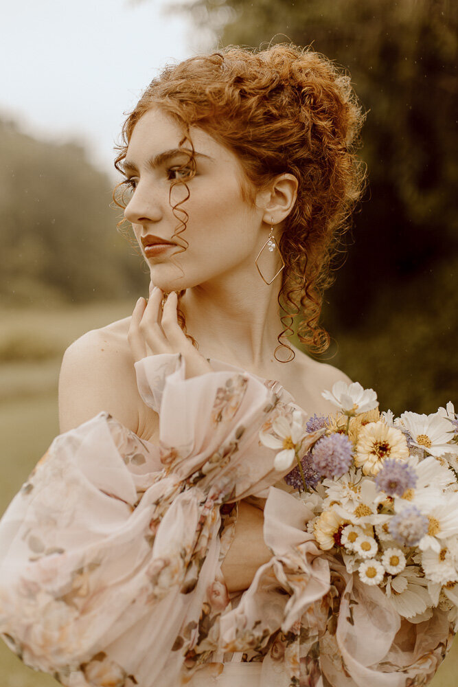 Bride poses in floral wedding dress in a a field while its raining