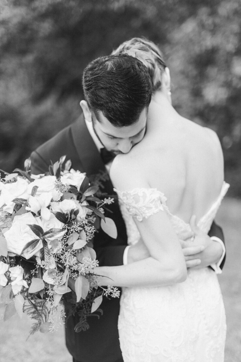 Black and white close-up of groom embracing bride with eyes closed, holding a large bouquet of roses and greenery.