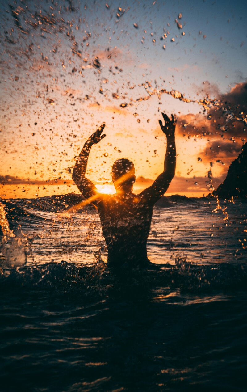 Image of a masc presenting person splashing in the waves at the beach at sunset, making a victorious pose with arms in air