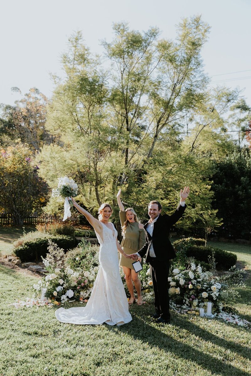 A couple celebrate their Central Coast Elopement with their Celebrant in front of a beautiful floral display and a native tree in the countryside location of Yarramalong. The couple are cheers and looking towards the camera. The image is warm and fun.