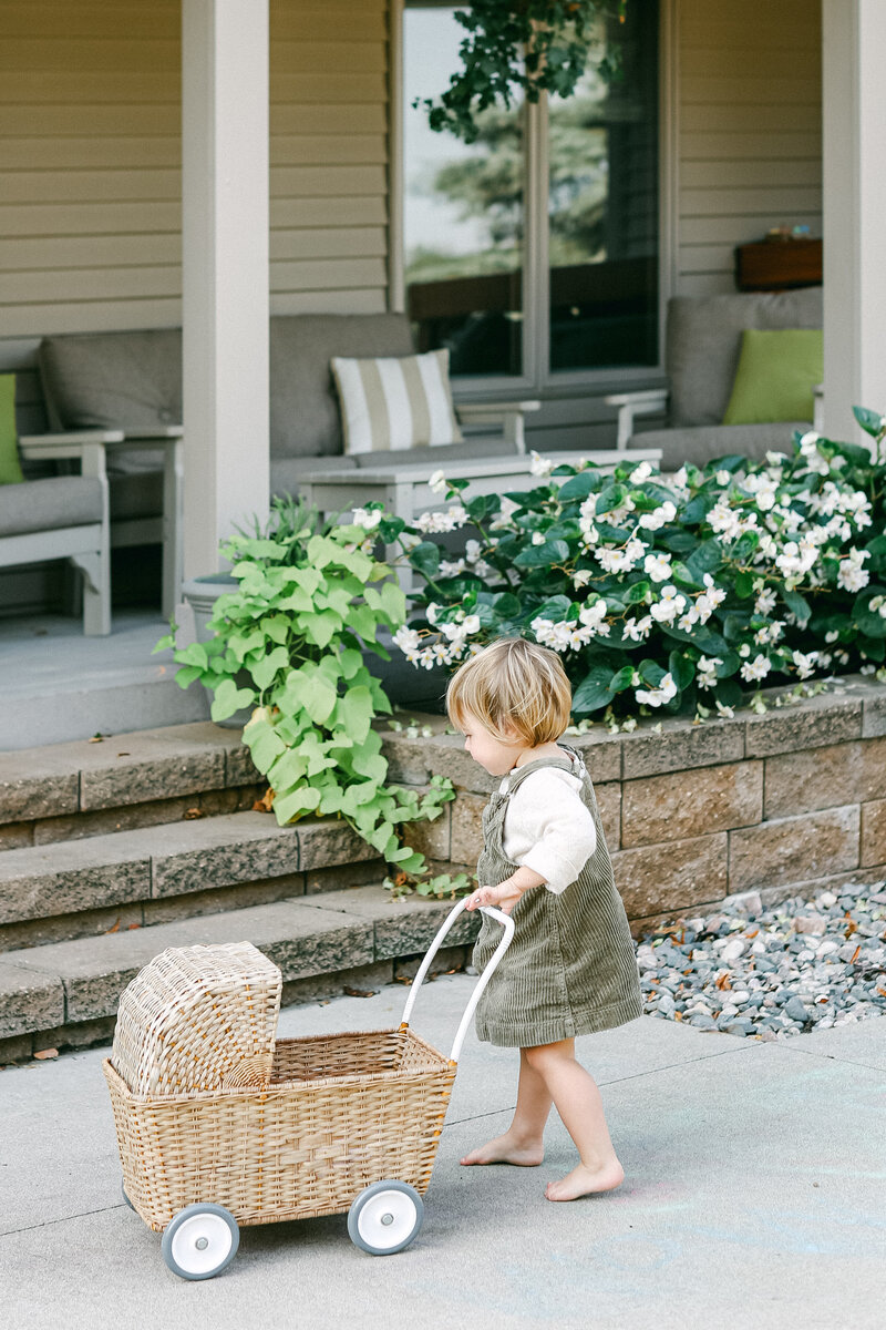 A toddler girl in bare feet pushing a wicker toy buggy near her front porch steps