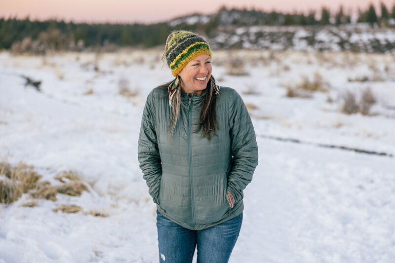 A woman wearing a green and yellow beanie and blue jacket laughs looking away from the camera on a snow covered landscape.