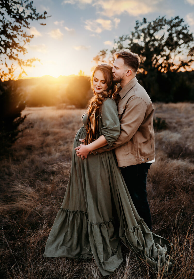 Maternity Photographer. a man kisses his pregnant wife as he embraces her from behind, she wears a dress with hands on her belly at golden hour outdoors