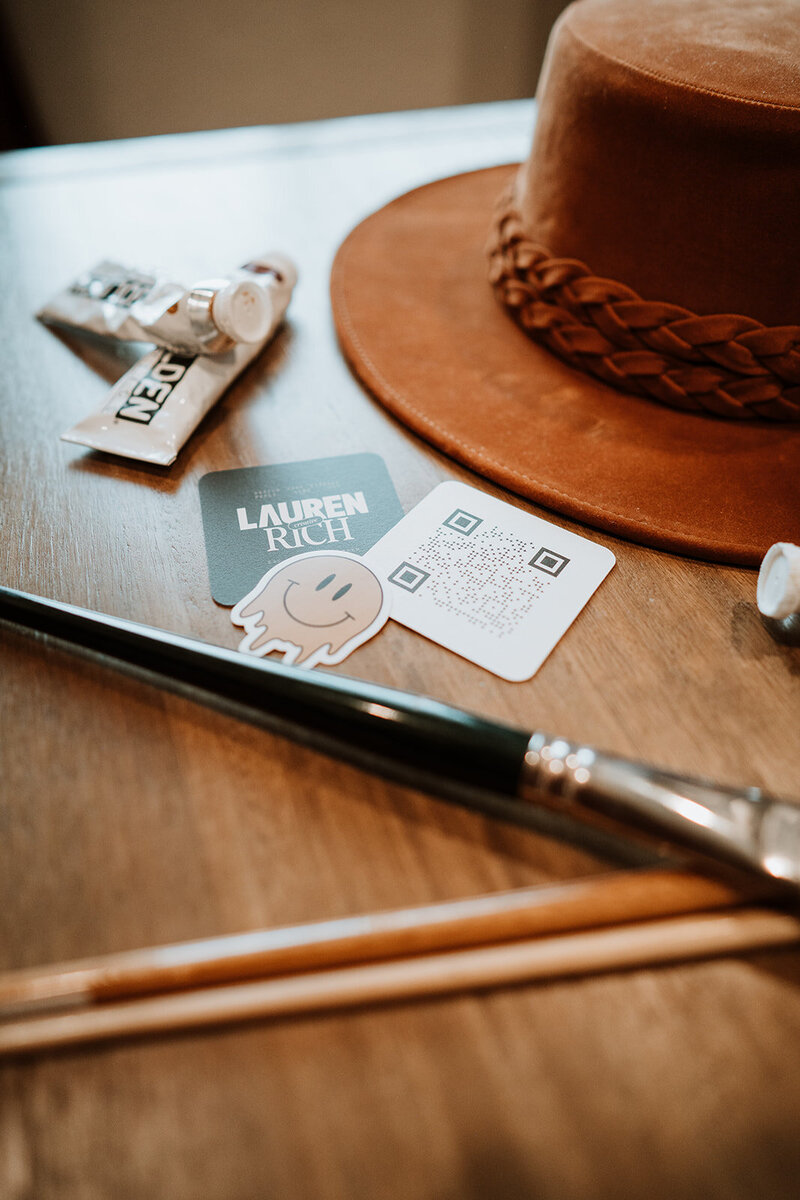 flat lay of hat, paint, brushes, business card, and sticker