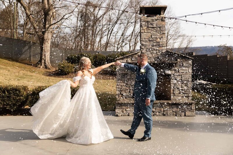 A newlywed couple dances outdoors at The Venue Chattanooga, surrounded by falling artificial snow. The bride, in an elegant lace ballgown, twirls gracefully while holding her groom’s hand. The groom, dressed in a tailored blue suit, smiles at her under the venue’s twinkling string lights, with a cozy stone fireplace in the background