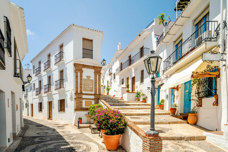 A bright, sunlit street in a Mediterranean-style village with whitewashed buildings, wrought-iron balconies, and terracotta flower pots. A stone staircase leads upward between the buildings, lined with plants and vibrant flowers. A classic black streetlamp stands in the foreground against a clear blue sky.
