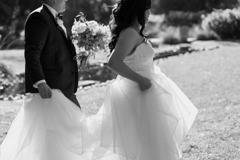 groom holds brides bouquet and wedding dress walking together