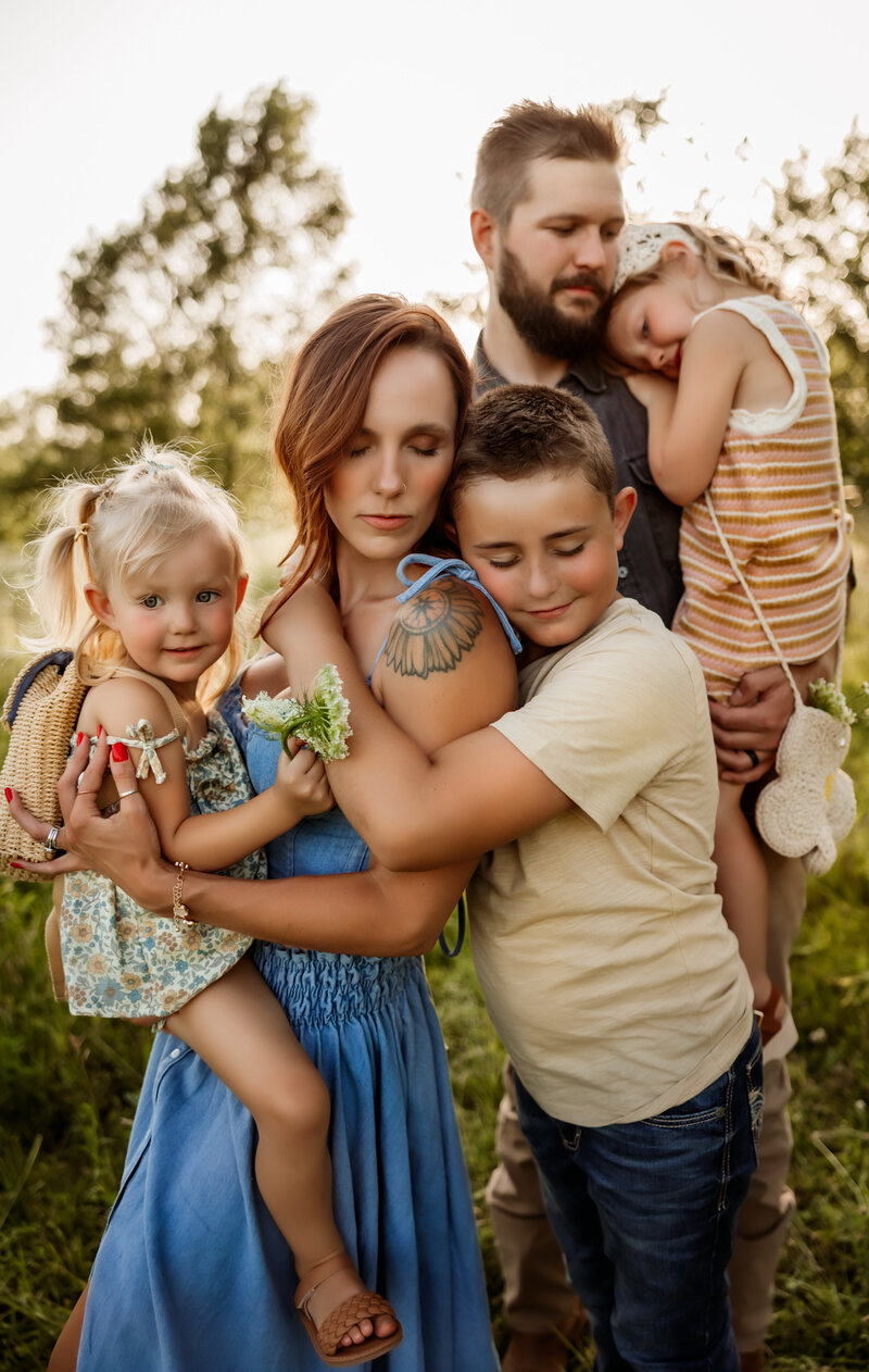 Mom holds little girl while son snuggles her with dad in the background holding their other daughter during family session with Olathe photographer.