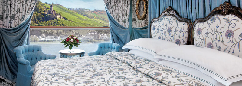 Elegant hotel or cruise-ship stateroom with ornate bedding, blue upholstered chairs, and large window overlooking a scenic riverside landscape.