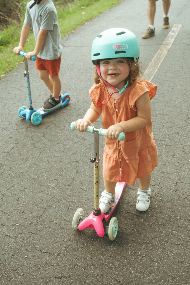 A cute toddler girl in a bright blue helmet riding a hot pink scooter.