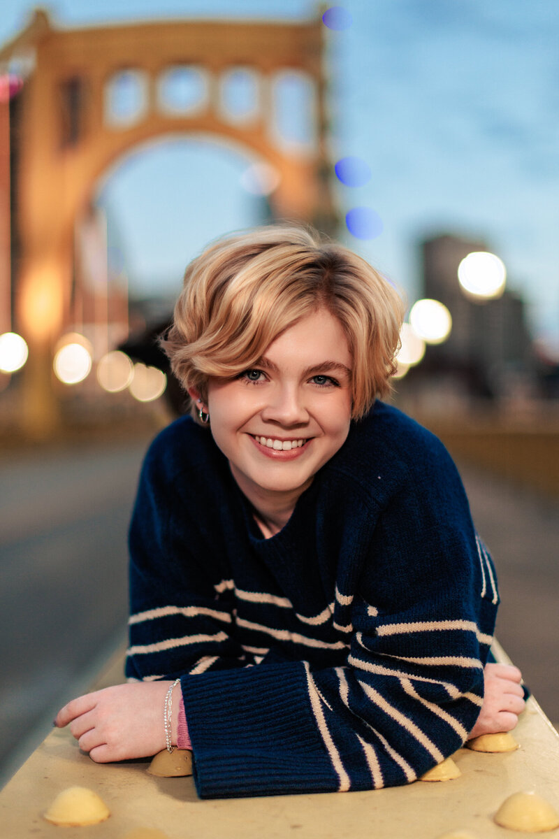 Graduating senior poses for pictures on the yellow pittsburgh bridge with the city lights behind her.