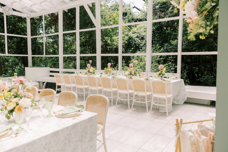 Long rectangular tables with floral runners and white chairs inside a bright glasshouse venue surrounded by lush greenery.
