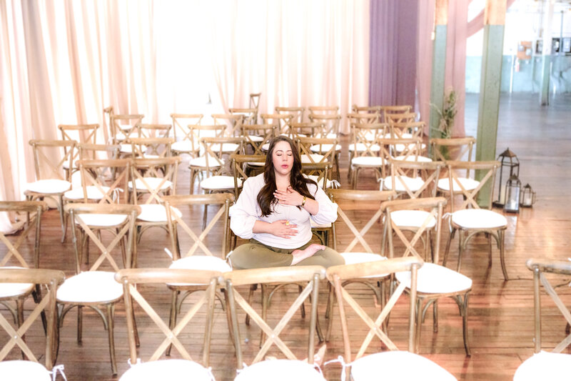Brunette girl with hands on heart and belly meditating in am empty room full of xback chairs