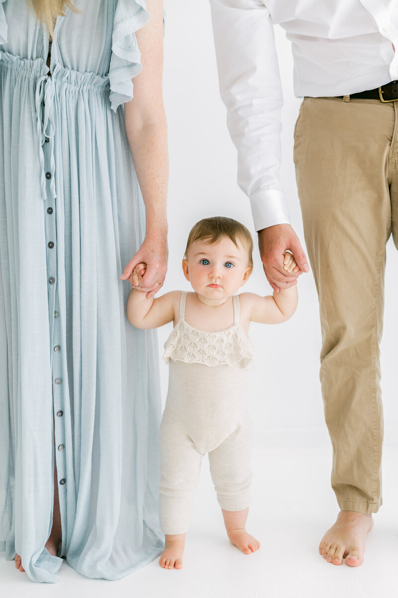 Baby girl looking at camera with neutral expression while mum and dad hold each of her hands