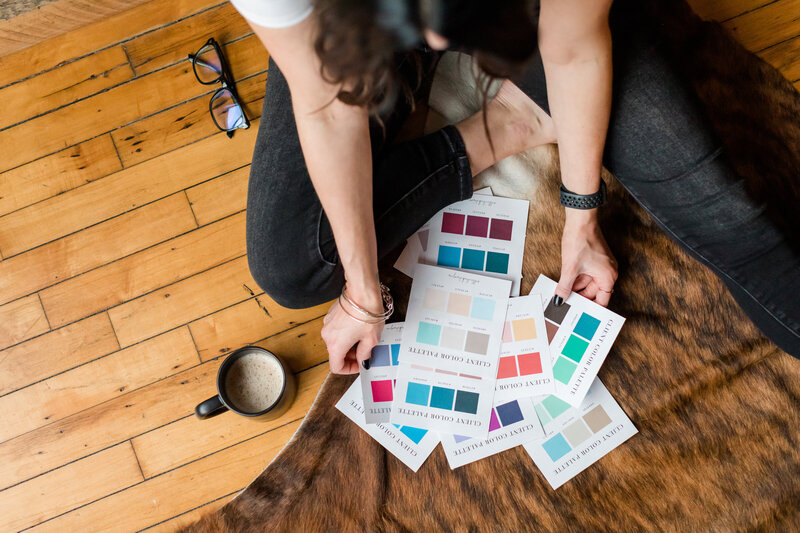 Overhead view of Allie Clark reviewing printed color palettes on a cowhide rug, surrounded by creative tools like a coffee mug and glasses — a behind-the-scenes glimpse into the brand development process at Allie Clark Designs.