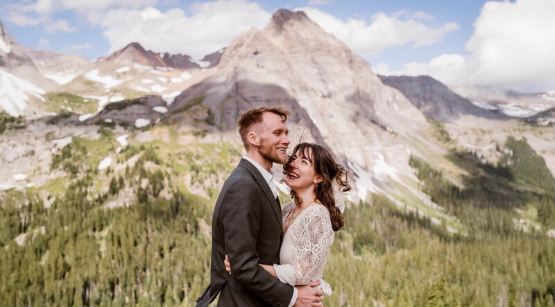 Bride and groom laughing together with dramatic rocky peaks and alpine forest in the background during their August elopement at Blue Lakes near Ridgway, Colorado, captured by Colorado elopement photographer Alpine Vows