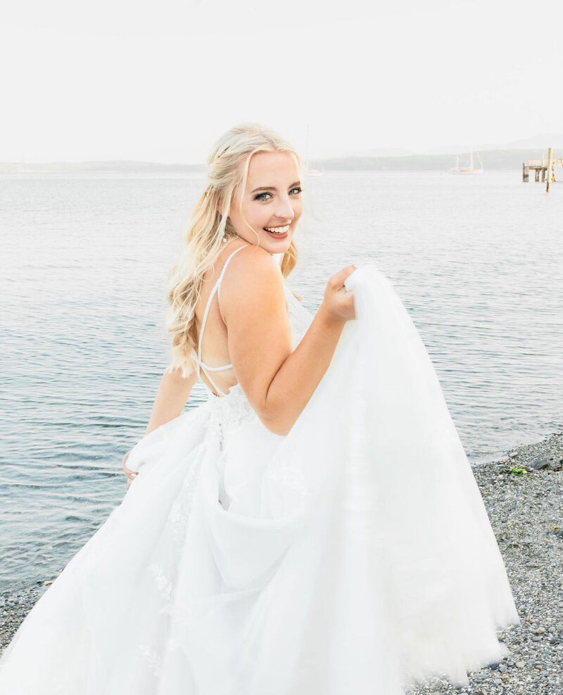 A bride standing by the water, holding her dress with a bright smile