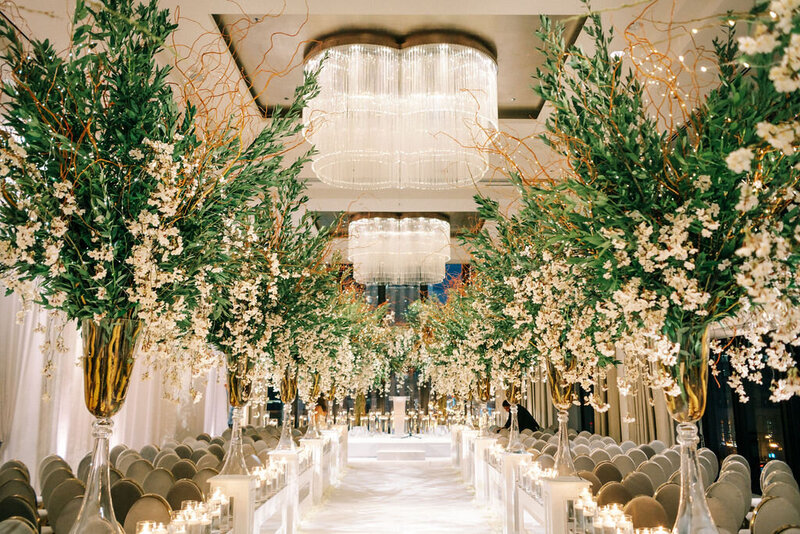 Magnificent wedding reception hall at The Langham Chicago, viewed from the back of the room, showcasing rows of elegantly set tables leading up to a floral-covered stage under a modern glass chandelier.