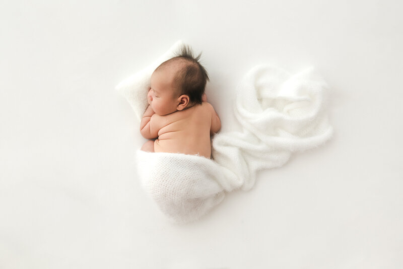 A newborn baby sleeps on a soft white blanket, curled up in a peaceful position. The blanket surrounds the baby, creating a gentle and serene atmosphere. The background is a smooth, solid white.
