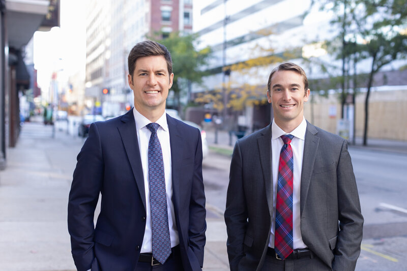 Professional headshots in Cincinnati: Two confident businessmen stand side by side against a city backdrop. The gentleman on the left sports a navy suit paired with a detailed tie, radiating leadership with his firm expression. To the right, his colleague in a gray suit and contrasting red patterned tie beams with a friendly smile, embodying approachability. Both exude professionalism in their tailored attire and poised demeanor