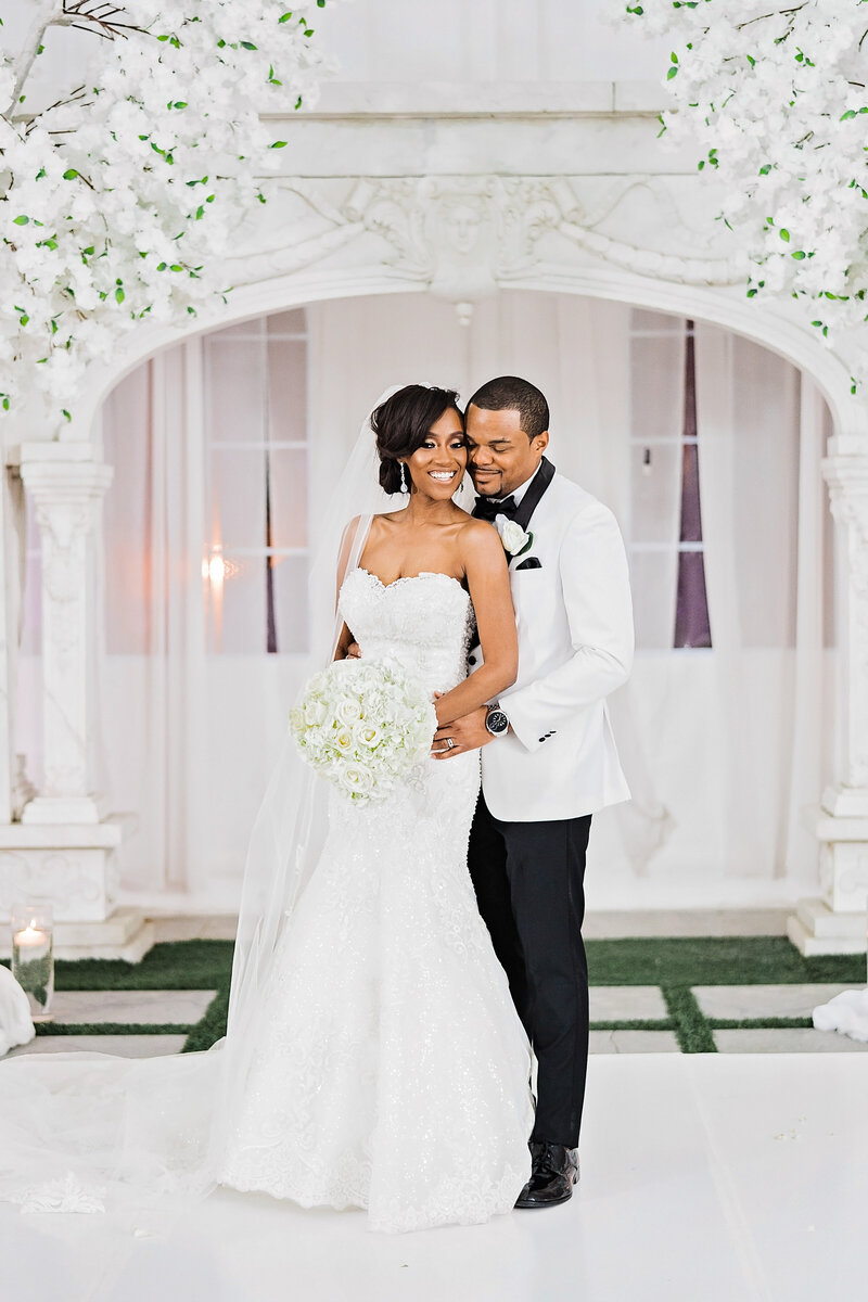 Groom hugs his wife from behind in a white room with an elegant arch behind them.