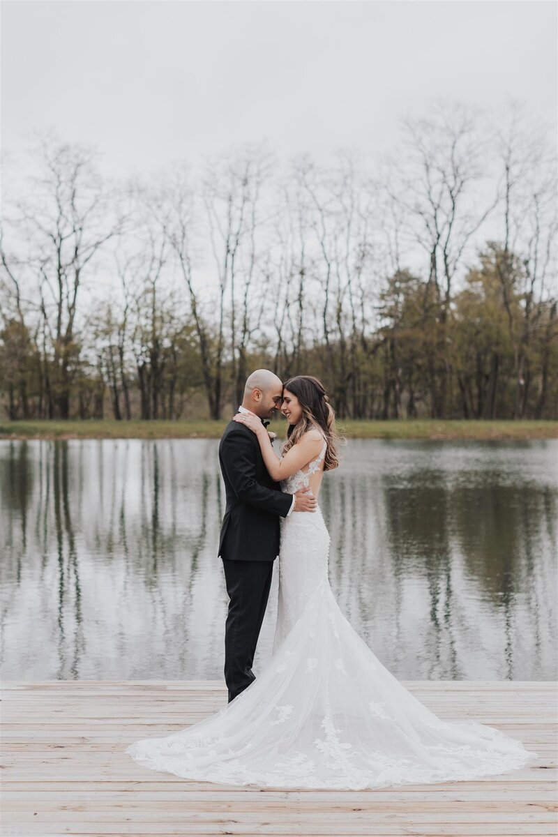 Classic groom and bride embrace as they stand on wood dock overlooking lake and trees in the Catskills