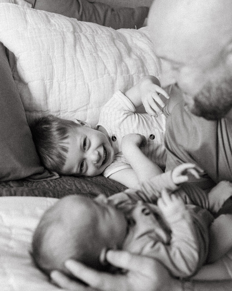 Toddler smiling directly at the camera with father and newborn baby slightly blurred in the foreground during a family photo session